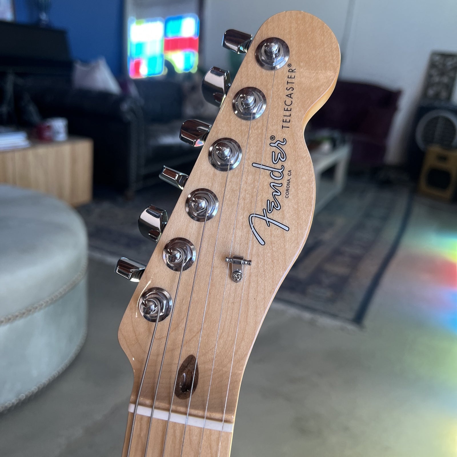 Close-up of a Fender Telecaster electric guitar headstock made of natural maple wood, showing six chrome tuning pegs and the Fender logo, with a blurred living room background.