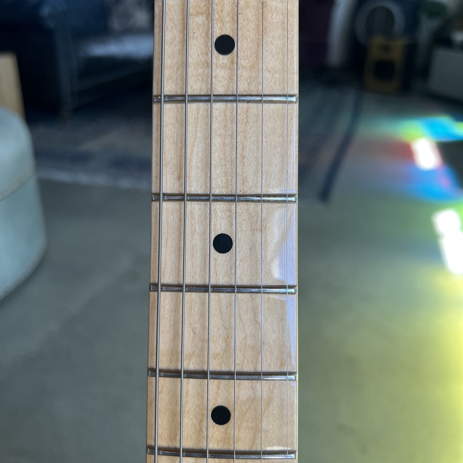 Close-up of a guitar neck fretboard with a light natural wood finish, metal frets, and black dot inlays, photographed indoors with a blurred background.