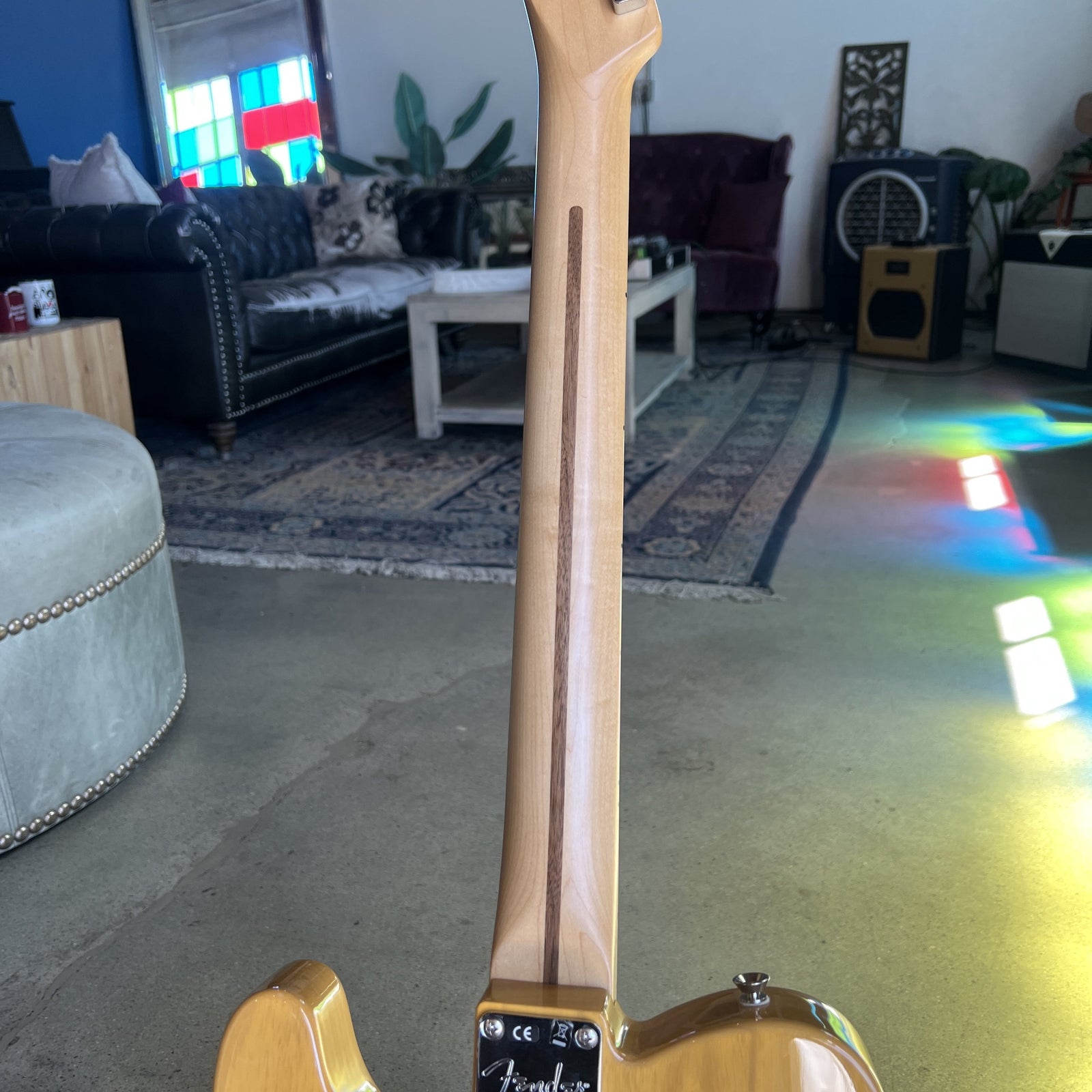 Close-up of the neck and lower body of a Fender electric guitar with a natural wood finish, photographed in a living room setting.