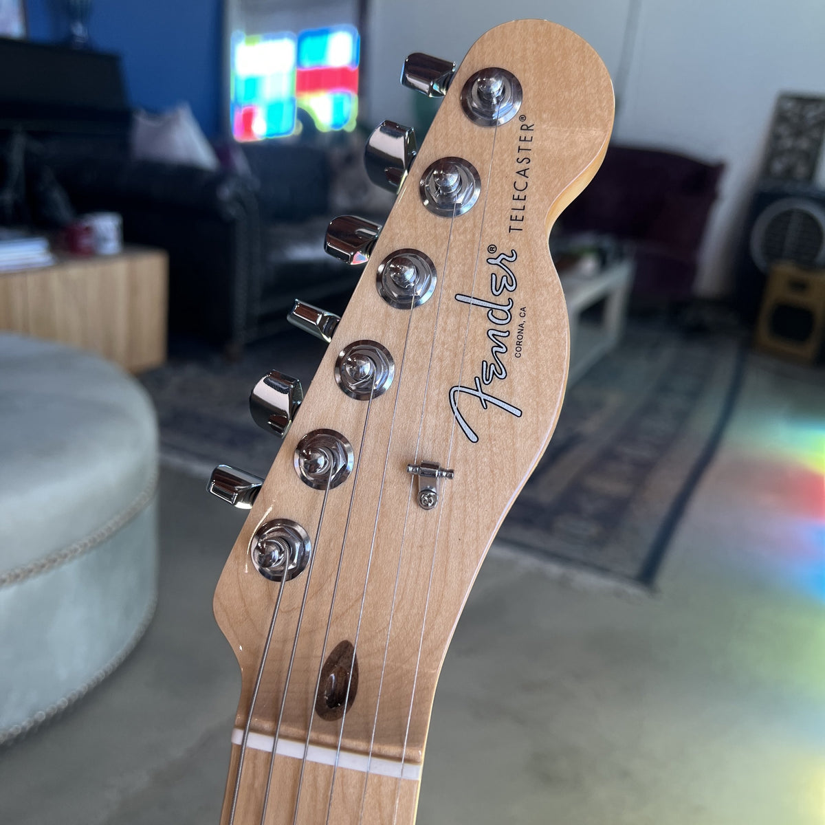 Close-up of a Fender Telecaster electric guitar headstock made of natural maple wood, showing six chrome tuning pegs and the Fender logo, with a blurred living room background.
