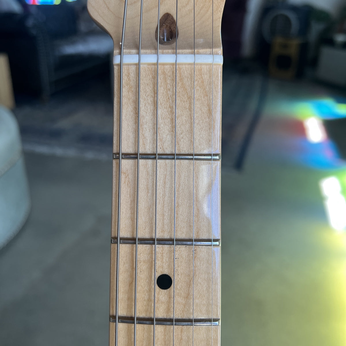 Close-up of a light natural wood guitar neck with six strings, metal frets, and a black circular fingerboard dot, photographed indoors with a blurred living room background.