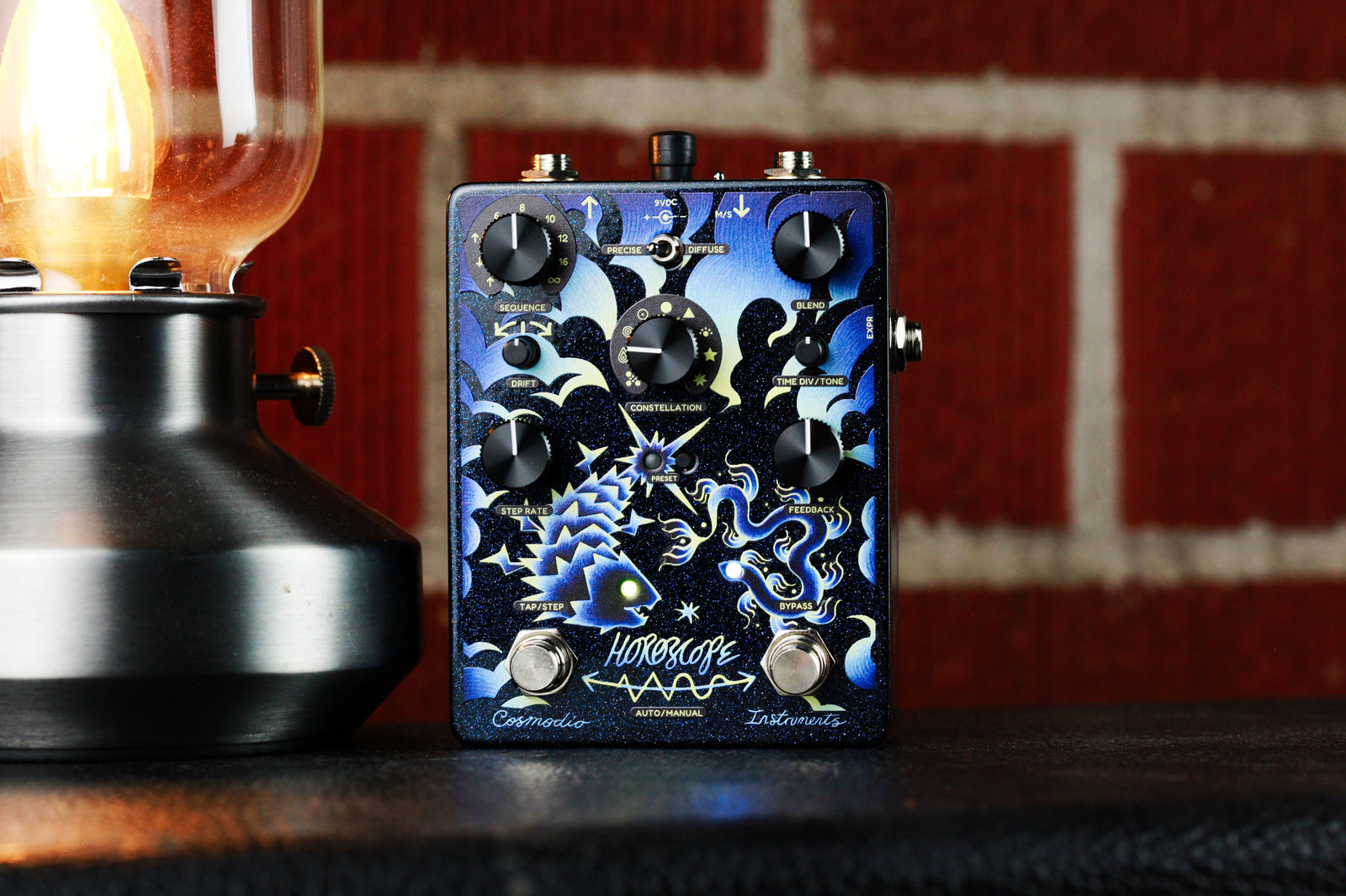 Cosmodio Instruments Horoscope Echo Sequencer pedal powered on with illuminated LEDs, displayed next to a warm vintage lamp against a red brick wall.   