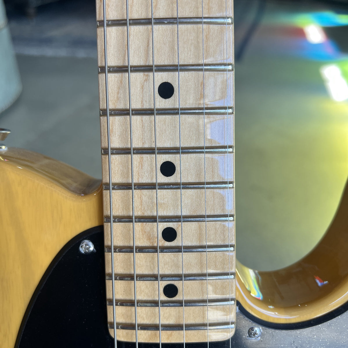 Close-up of an electric guitar's neck, showing a light natural wood fretboard with black dot inlays and a glossy yellow-orange body.