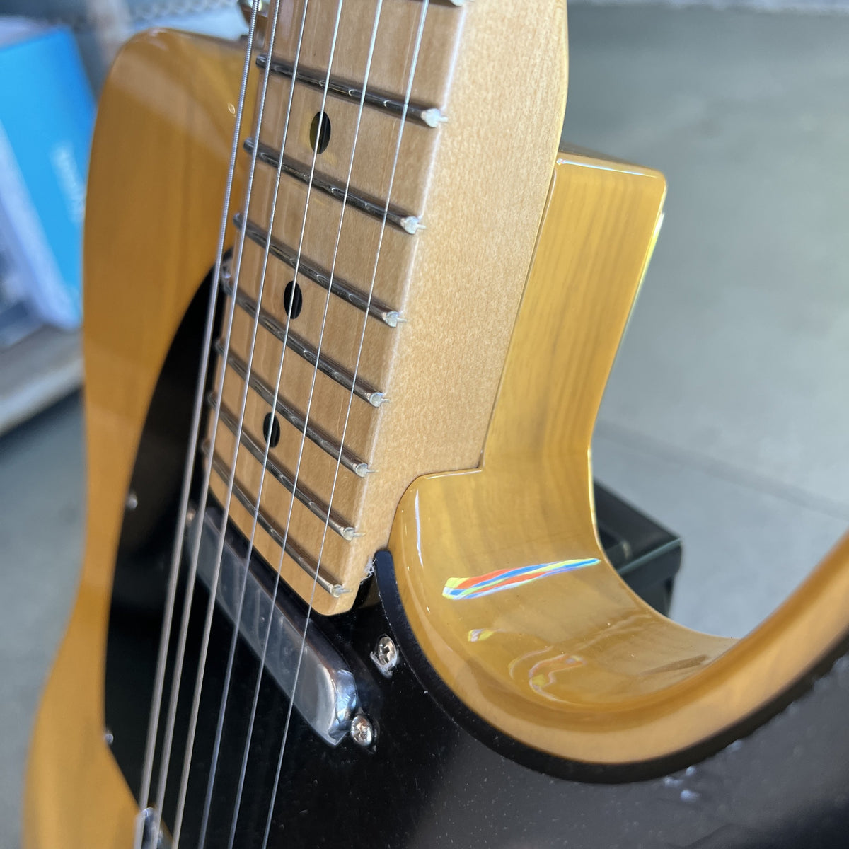 Close-up of a T-style electric guitar body with a glossy gold finish, black pickguard, and part of the fretboard visible, photographed indoors with a blurred background.