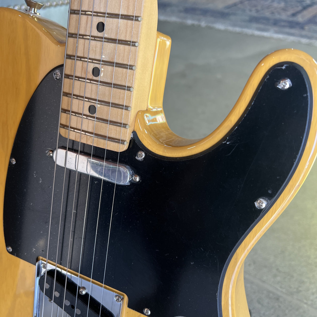Close-up of a classic electric guitar featuring a natural maple wood body, black pickguard, and maple fretboard with black dot inlays.