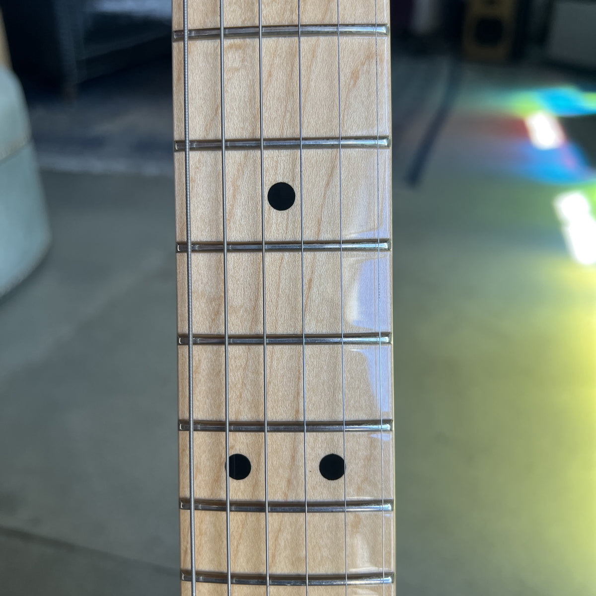 Close-up of a light natural wood guitar fretboard with metal frets and black dot inlays, photographed indoors with a blurred background.